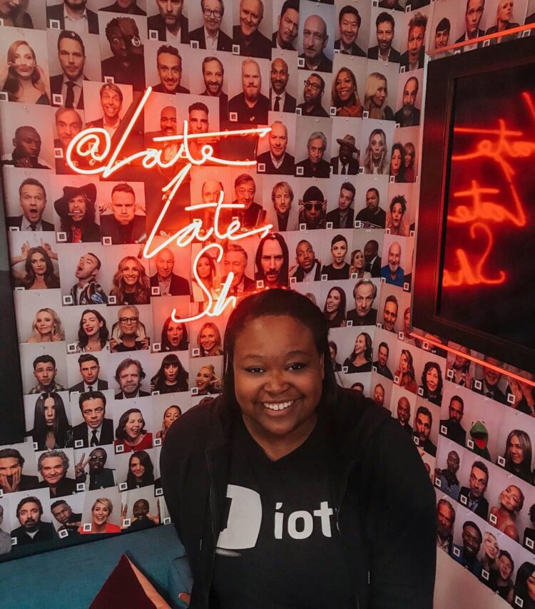 Sha Collier poses in front of a wall full of headshots with a neon sign that says 