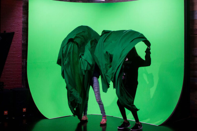 Two girls hide behind green sheets while standing in front of a green screen.